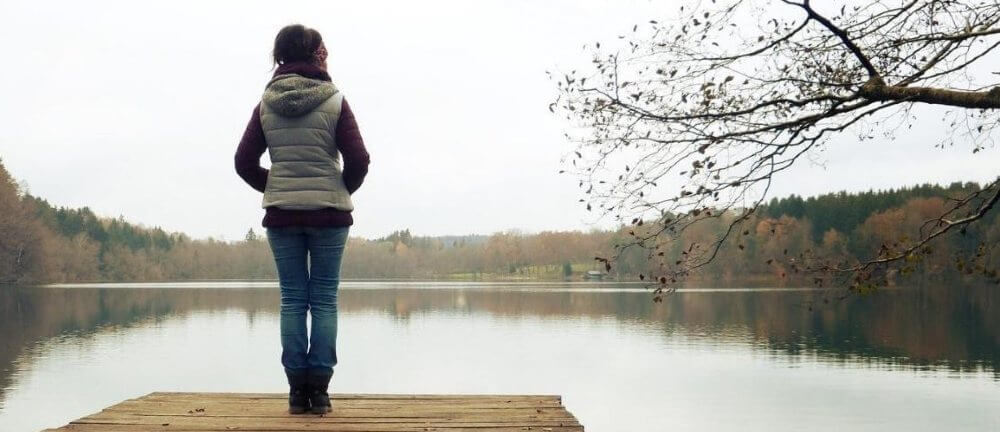 Missbruk, trauma, samsjuklighet. En kvinna står på en brygga och blickar ut över en sjö. Vädret är grått och dystert.