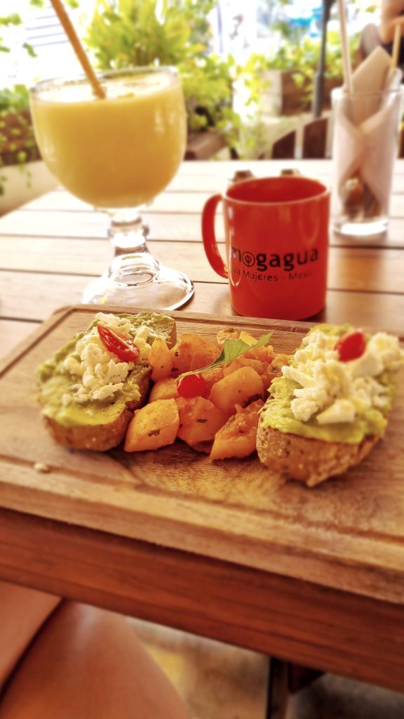 Cafe Mogagua is one of the best places to visit for breakfast on the island. This photo shows a Cafe Mogagua mug with a big glass of juice and avocado and feta on toast.