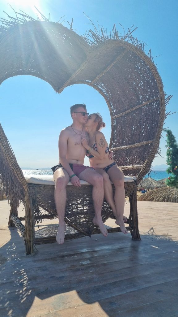 Amy and Liam sitting on a thatched love heart seat on Paradise Island. Amy is leaning into Liam kissing his cheek with the sun beaming down on them.