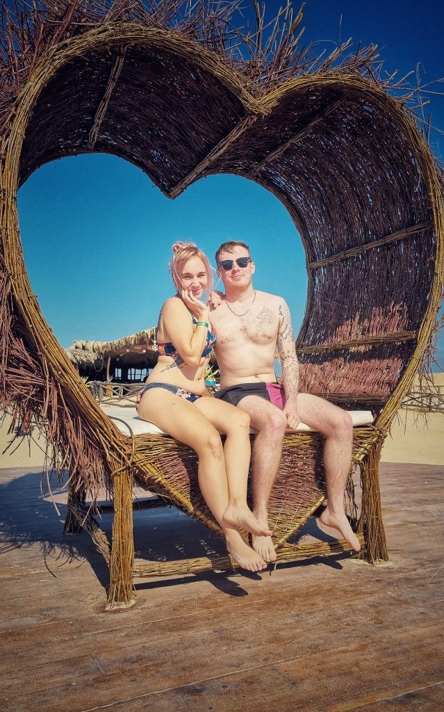 Amy & Liam sitting on a thatched love heart seat together.