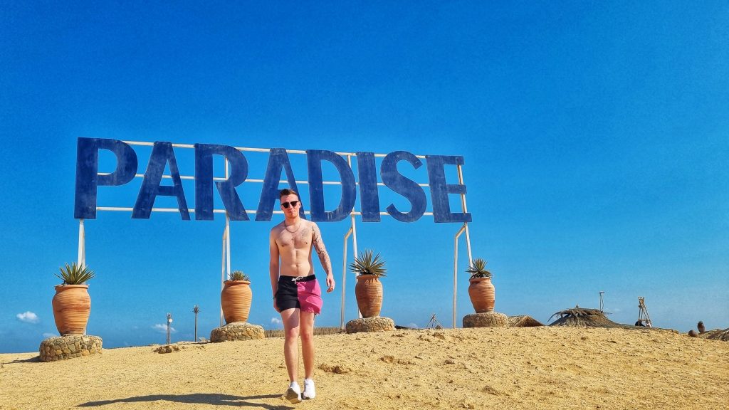 Paradise Island sign in large blue letters with Liam walking away from the sign towards the camera.
