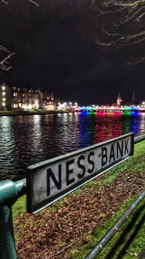 Inverness Bridge is lit up in the distance with different colours. At the forefront of the photo is a sign for Ness Bank.
