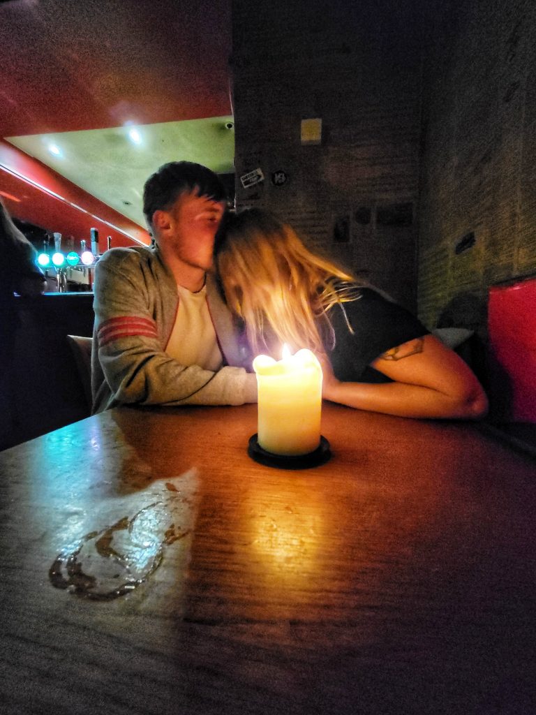 Amy and Liam enjoying their time at Hootenannys. This image shows Amy leaning into Liam and he is kissing her on the head with a candle lit in front of them.