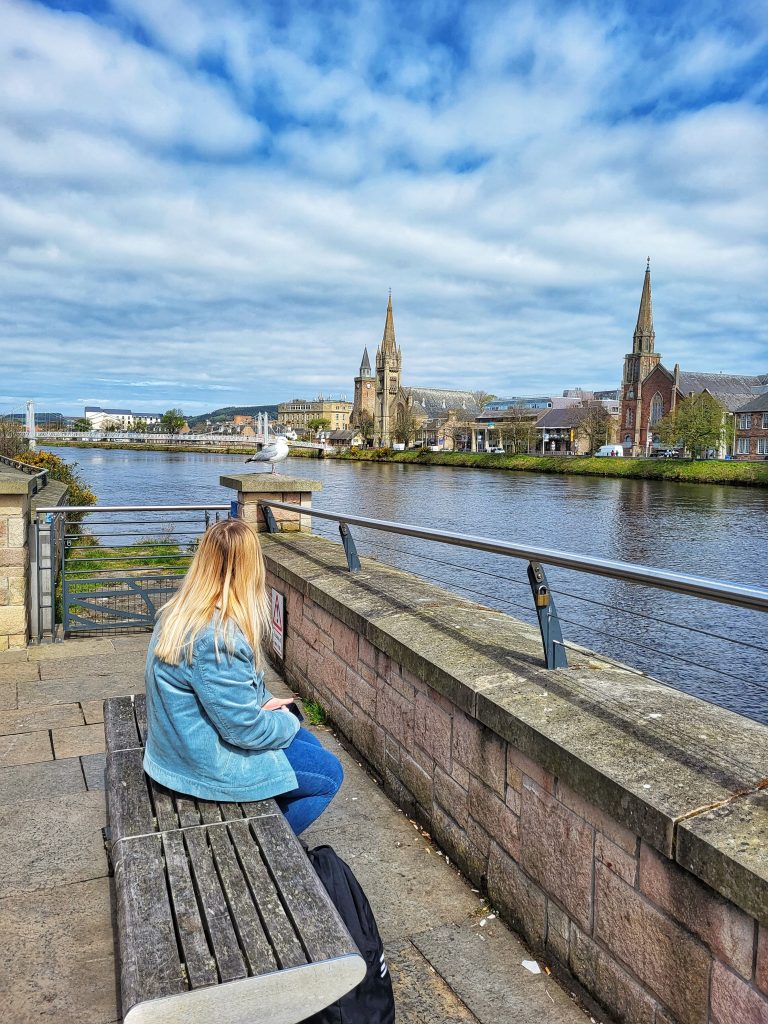 Amy sitting on a bench at Ness Bank where you can see the cathedral over the other side.