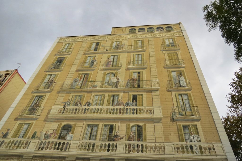 Some of the beautiful street art in Barcelona on the side of a building, It shows different residents standing on the balconies. This street art is clever as the actual building doesn't have any windows.