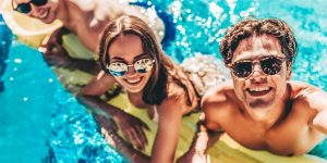 Happy attractive young friends in sunglasses are having fun in swimming pool on an inflatable mattress and smiling while making selfie.