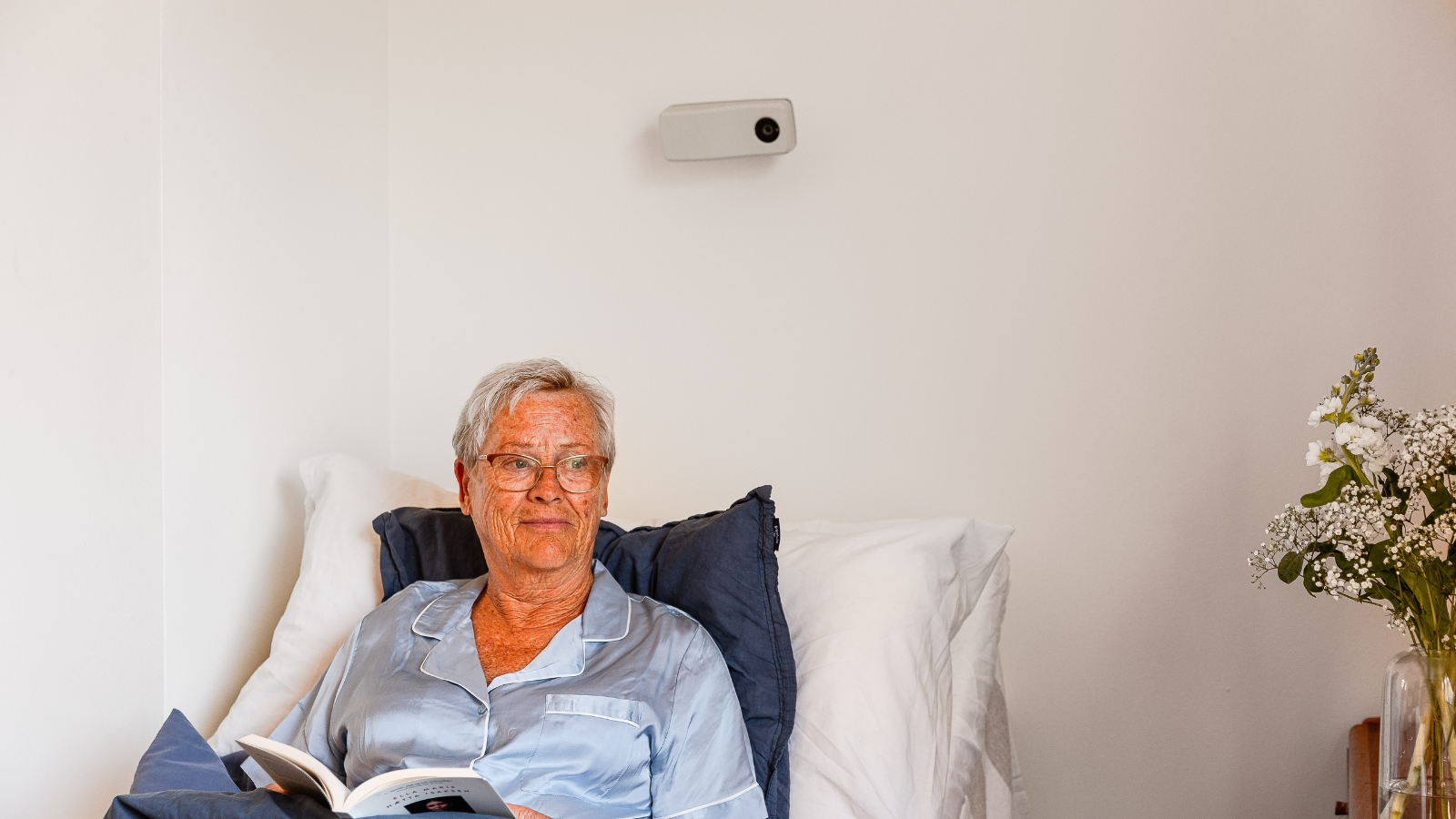 A woman resting in bed with a digital supervision device (Night Owl) above on the wall.