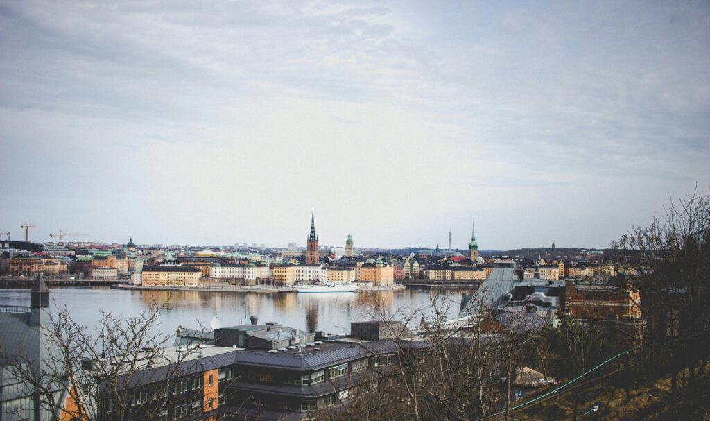 En panoramavy över en stad med en stor vattenmassa i förgrunden. Stadens silhuett domineras av kyrktorn och färgglada byggnader, och spegelbilden av byggnaderna syns på vattenytan. I bakgrunden sträcker sig staden vidare under en molntäckt himmel.