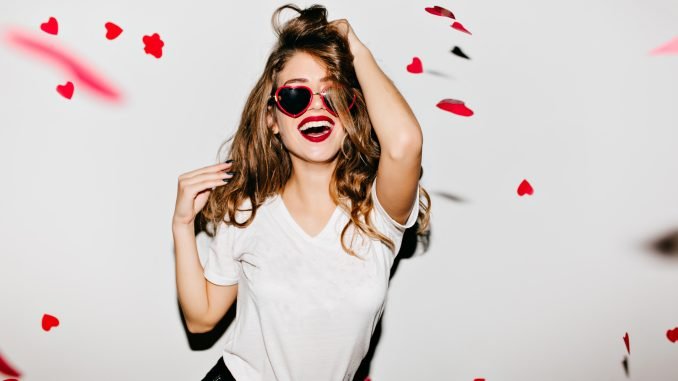 Photo by Aiko Guszman Indoor portrait of amazing caucasian female model in trendy t-shirt touching her long shiny hair. Laughing refined woman in sunglasses having fun with red confetti.