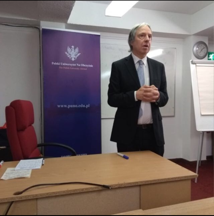 A man is standing behind a desk, talking to a classroom