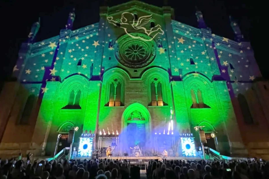 Piazza del Carmine a Pavia di notte durante un concerto del Festival del Carmine. La storica facciata della chiesa trecentesca è interamente illuminata da proiezioni di luci verdi e gialle con stelle e un angelo. Sul palco rialzato si esibisce una band davanti a una folla di persone sedute in platea.