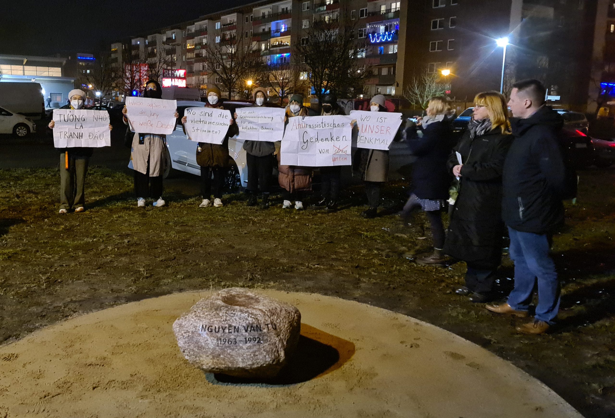 Es ist dunkel, der Ort wird nur durch Straßenlaternen beleuchtet. Im Vordergrund liegt ein Findling mit der Aufschrift Nguyen Van Tu 1963 - 1992 auf einer Fläche aus hellem Sand, die sich auf einer Wiese vor dem Parkplatz des Einkaufszentrums befindet. Dahinter stehen 6 Leute , die Mützen und Coronamasken tragen und halten weiße Papiere mit handgeschriebenen Aufschriften wie: warum saßen in der Jury nur weiße Deutsche, wo sind die vietnamesischen Stimmen, Für Euch Gewissenserleichterung für uns kollektiver Schmerz, Antirassistisches Gedenken von unten oder wo ist unser Denkmal, hoch. Eine der Aufschriften war auf vietnamesisch.