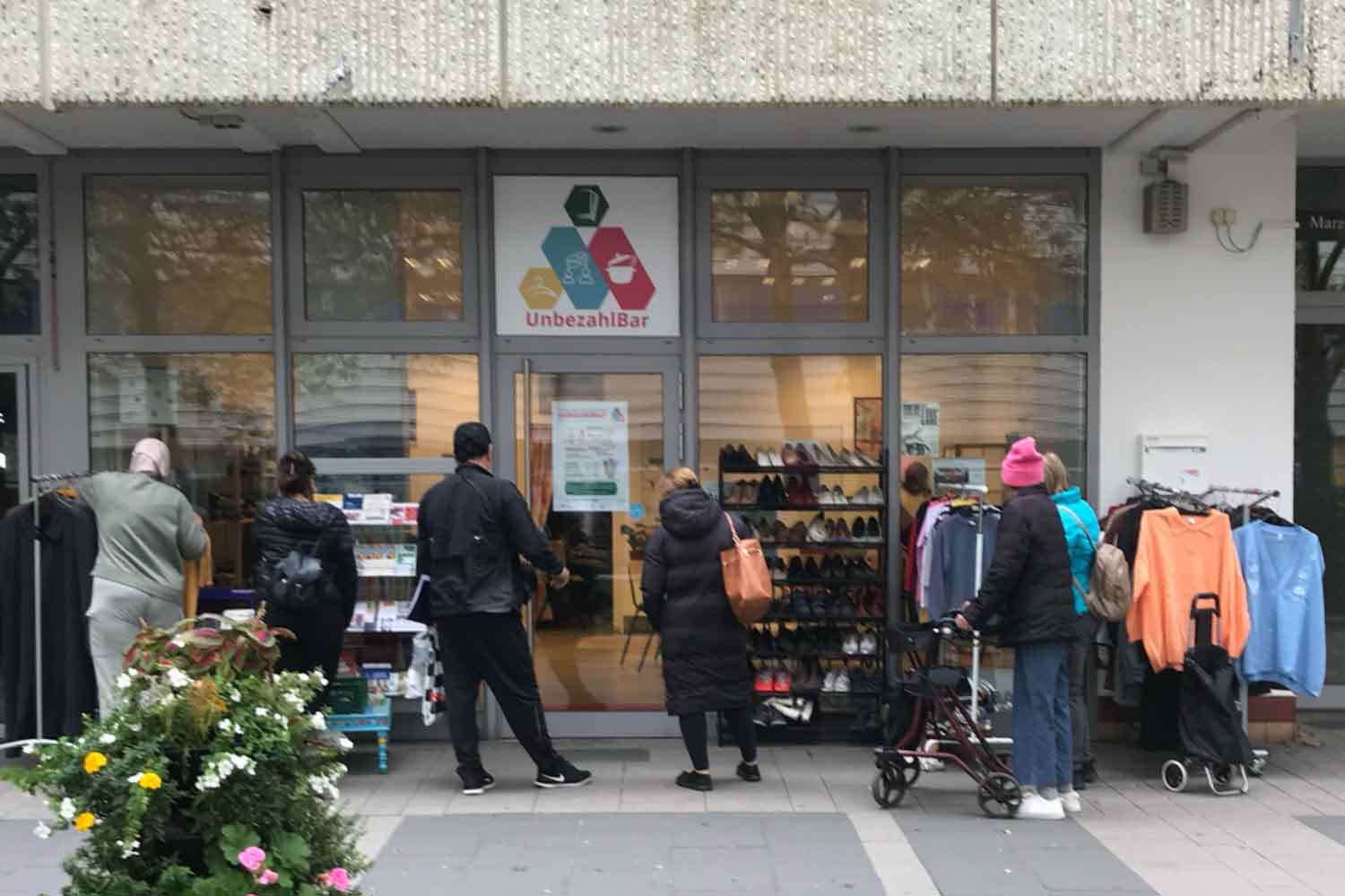 Vor einem Laden stehen 6 Leute und und gucken sich Schuhe und Kleidung an den Kleiderständern davor an. Über dem Laden hängt ein buntes Schild auf dem steht UnbezahlBar.