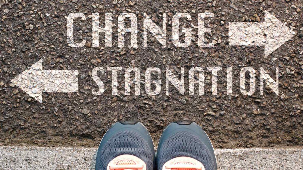 Billedet forestiller et par sko, der står på en vej med skriften 'change' pil til højre og 'stagnation' pil til venstre, som symbol for at stå ved en skillevej i livet.
