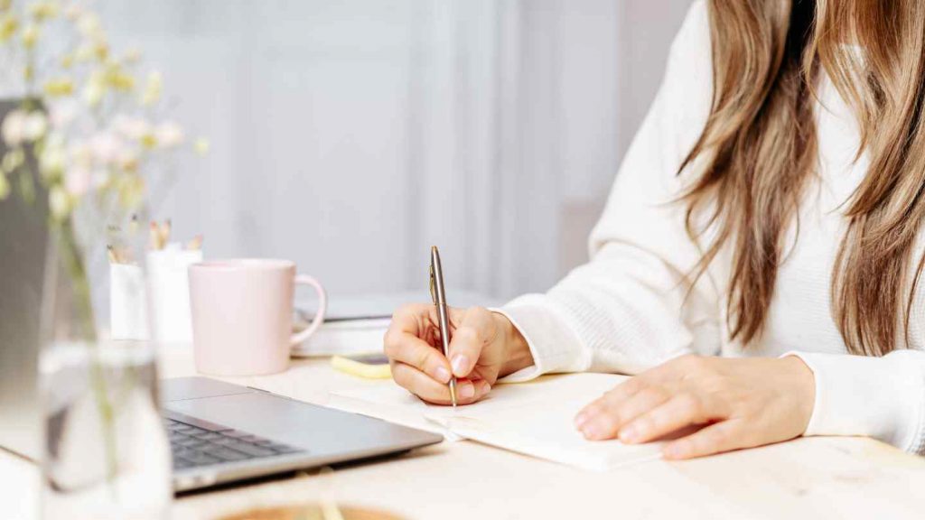 Billedet forestiller en kvinde, som sidder ved et skrivebord, hvor der står en kaffekop, en computer og foran hende ligger en blok. I hånden har hun en kuglepen og hun er ved at lave en øvelse for at finde hendes livsværdier.