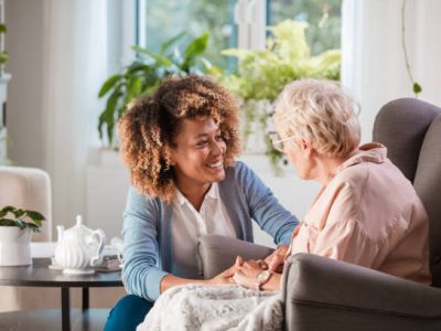 Female home caregiver talking with senior woman, sitting in living room and listening to her carefully.