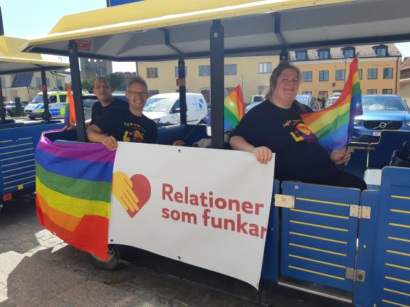 Petter, Magnus och Tess sitter i ett Pride-tåg. DE håller en Relationer som funkar-flagga och regnbågsflaggor.