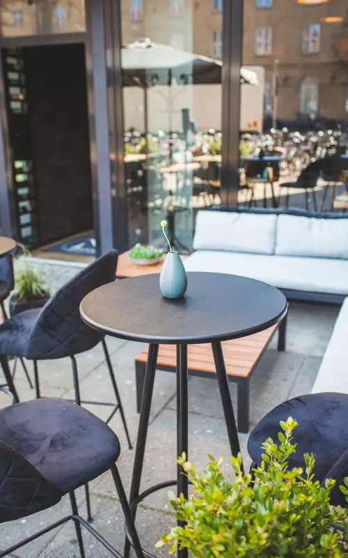 Et billede af udendørs loungemøbler på Restaurant Nögen i Aarhus. De behagelige sofaer og stole er placeret på en flisebelagt terrasse, der er omgivet af grønne planter og træer. Der er også en stor parasol, der skaber skygge og giver beskyttelse mod solen. En af sofaerne har bløde puder og en tæppe, som inviterer gæsterne til at slappe af og nyde den afslappende atmosfære på restaurantens udendørsområde
