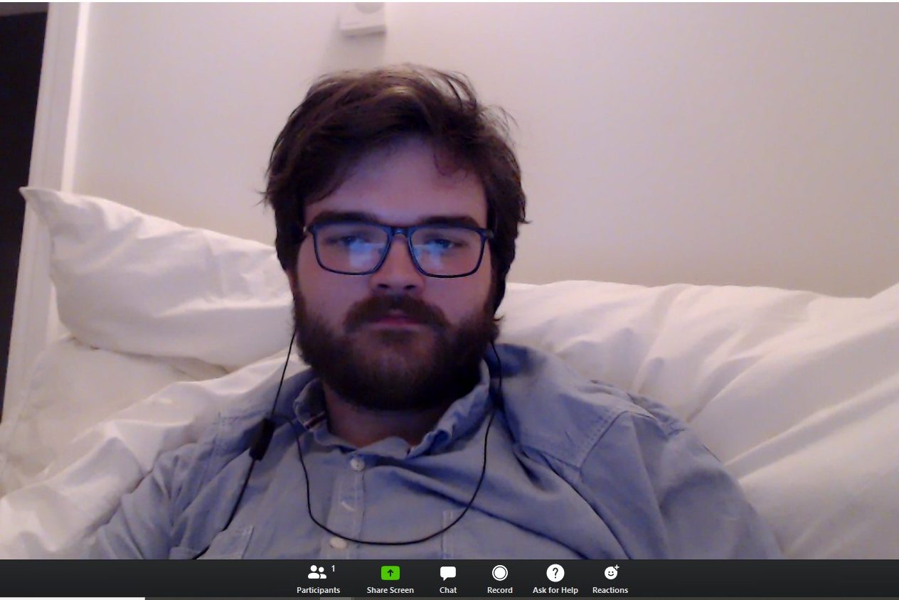 A young man, sits alone in a zoom call, looking dejected.