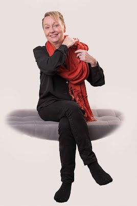 Monica af Klinteberg har svarta kläder och en orange halsduk, sitter på en grå soffa.