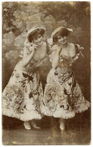 Old photo of two female dancers with gibson girl hair, wearing embroidered ankle length dresses with a deep neckline, leaning forward and raising one finger each
