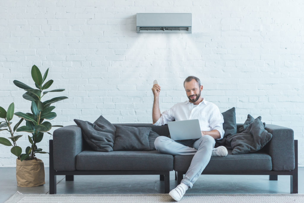 kjekk mann som slår på klimaanlegget med fjernkontroll mens han bruker laptop på sofaen hjemme