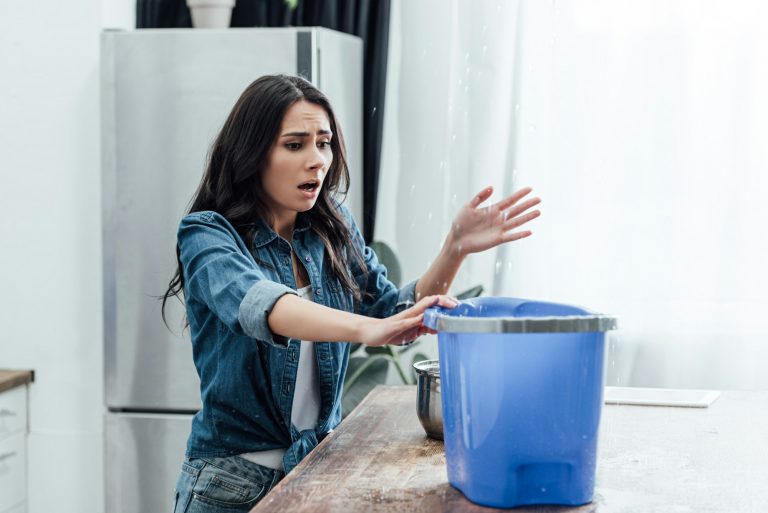 Upset young woman using bucket during leak in kitchen