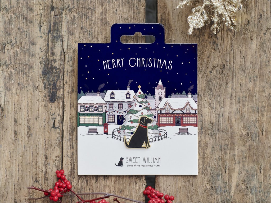 Image shows a Christmas gift item - a dog pin sitting on a packaging background showing a Christmas scene