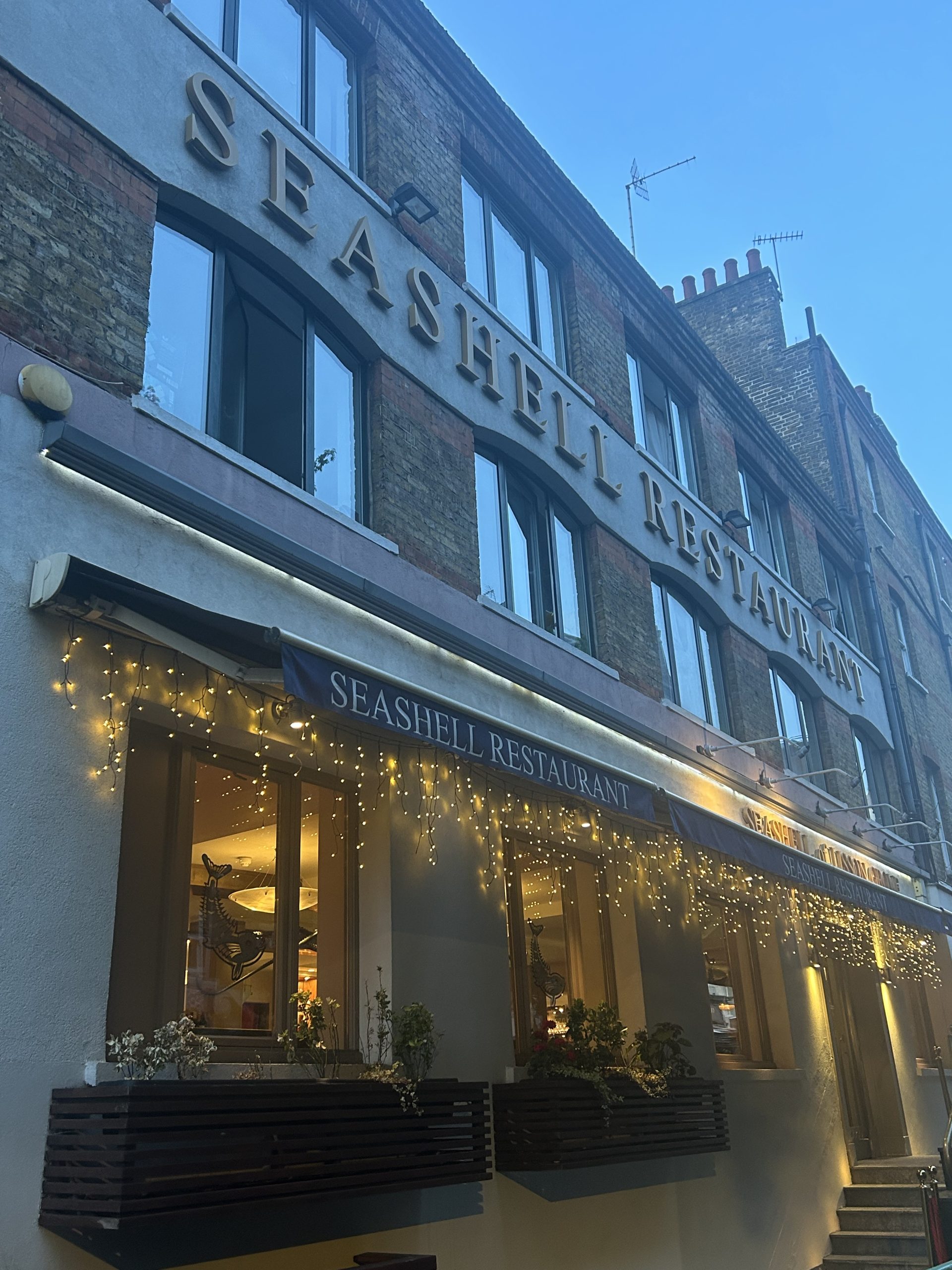 Exterior view of The Seashell Restaurant in London at dusk, featuring illuminated signage, warm fairy lights across the entrance, large windows and a classic brick building facade.