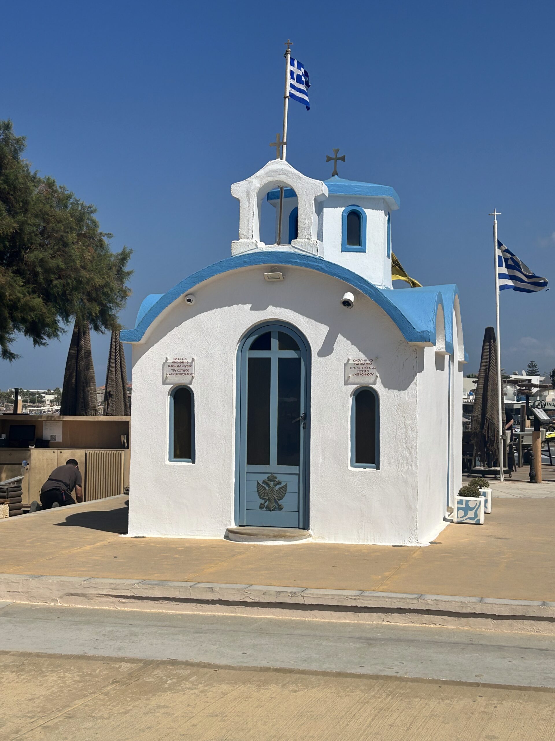 Small white and blue Greek Orthodox chapel in Crete with Greek flags flying against a clear blue sky.