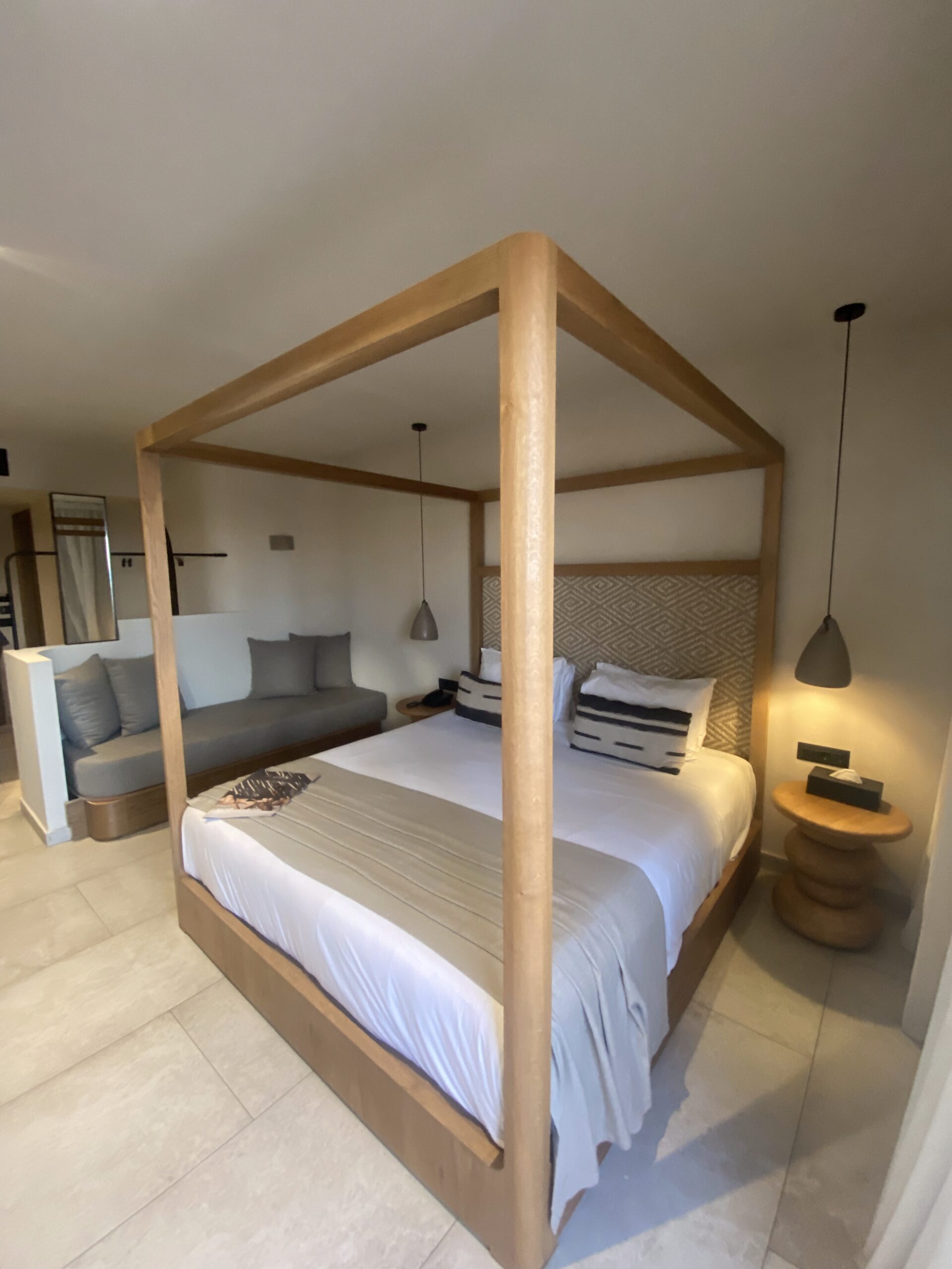 Elegant bedroom at Stella Blue Crete featuring a wooden four-poster bed, neutral earthy decor and a bright balcony overlooking palm trees.