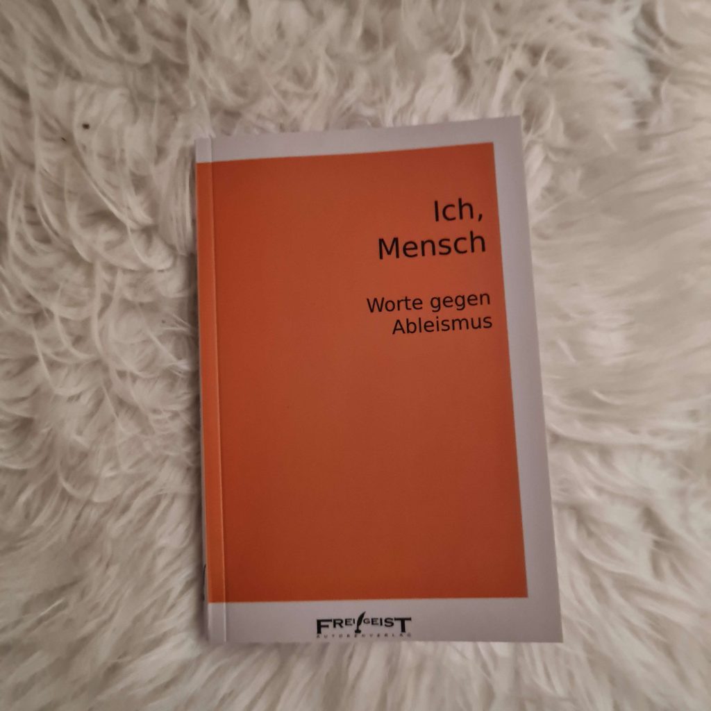 Ein Buch liegt auf einem Fell. Buchcover: Schwarze Schrift auf orangefarbenem Grund: Ich, Mensch Worte gegen Ableismus Frei!Geist Autorenkollektiv