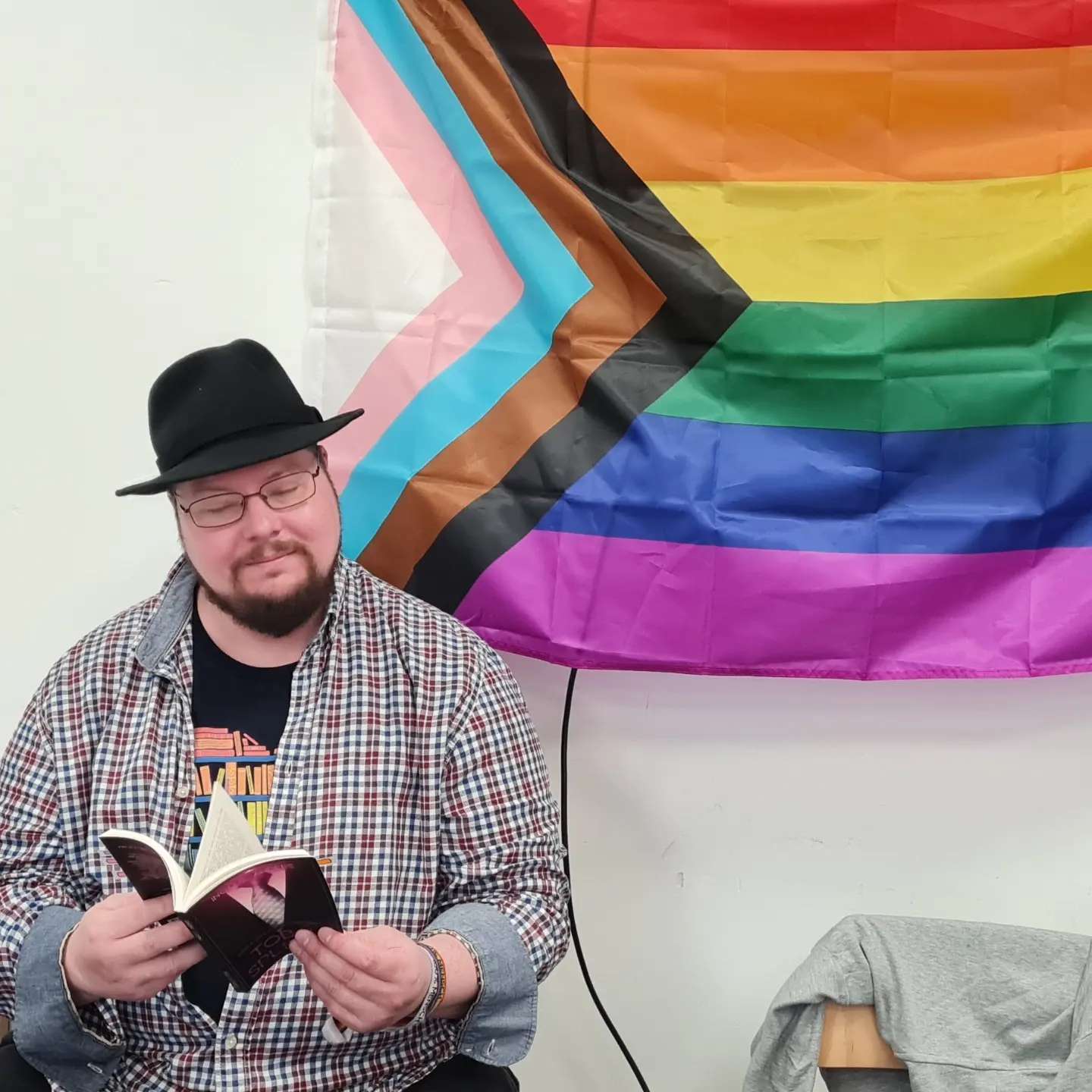 Ein Mann mit Hut liest aus einem Buch. Im Hintergrund die Progressive Regenbogen-Fahne.