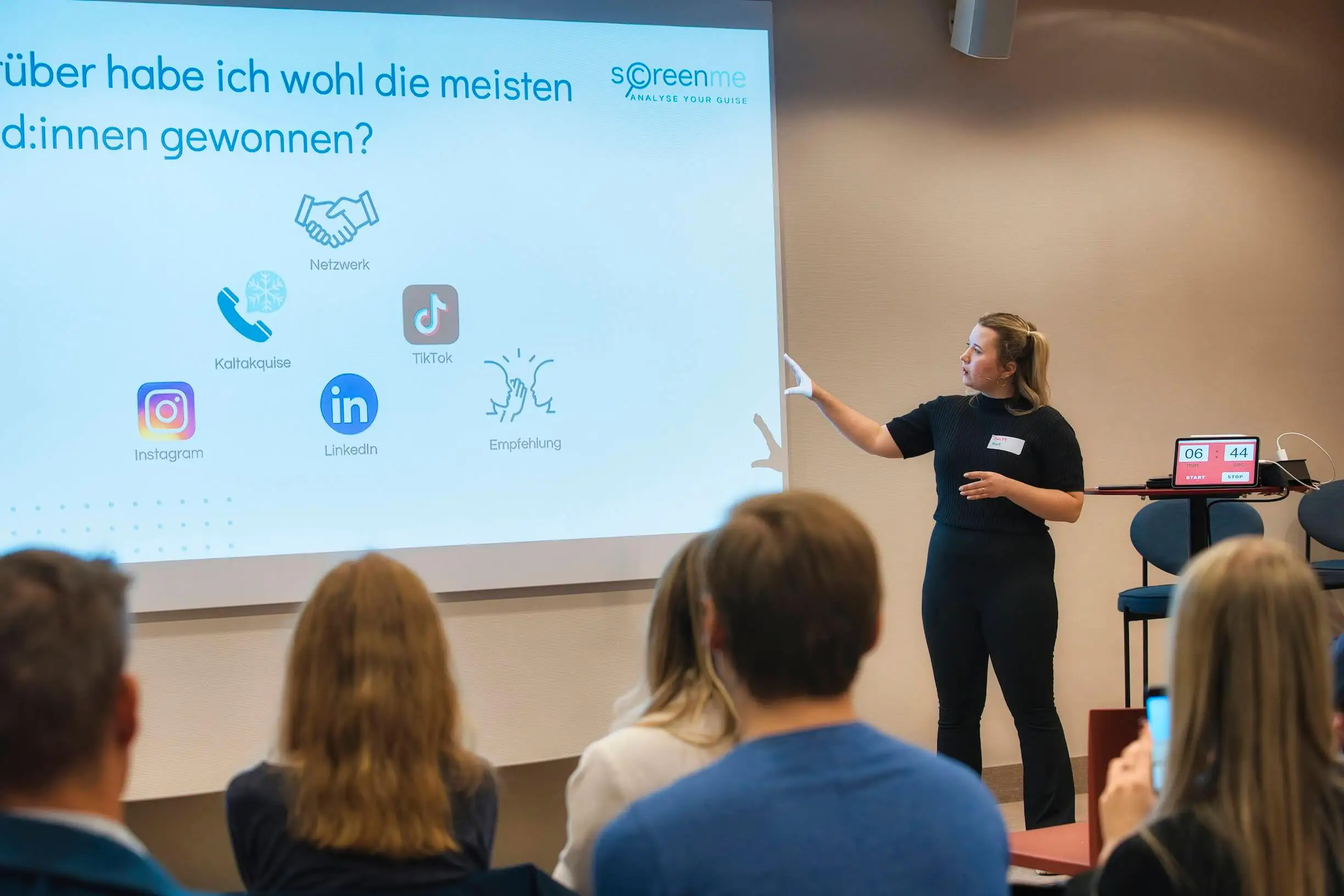 Konferenz mit Präsentationsfolien, Frau steht vor Publikum und hält einen Vortrag