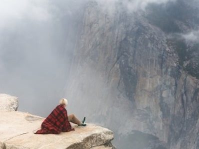 Eine Person, die eine rot-schwarz karierte Decke trägt, sitzt am Rand einer Felsklippe und blickt auf eine neblige Berglandschaft mit tief hängenden Wolken. Die Person scheint eine Flasche zu halten, und ein Seidenschal ist locker um ihren Hals geschlungen, was ihr einen Hauch deutscher Mode verleiht.