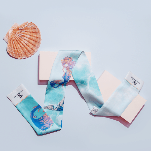 A Seidentuch "Meerjungfrau" scarf, featuring an illustration of a mermaid and ocean waves, is artistically draped atop a light pink rectangular box, with a seashell placed nearby on a light blue background.