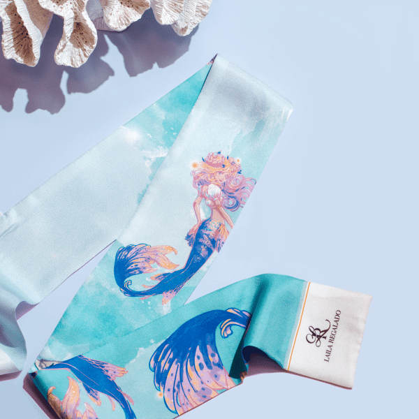 A silk scarf, Seidentuch "Meerjungfrau," with a colorful mermaid design is draped on a light blue surface. The scarf features shades of blue, pink, and turquoise, with an illustrated mermaid. A coral piece is partially visible in the background. A label shows "LALIA WORLD" branding.