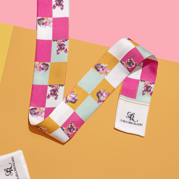 The Seidentuch "Frenchie" 115x4 is a luxurious silk scarf adorned with a vibrant checkered pattern featuring squares in pink, yellow, white, and turquoise, interspersed with charming dog illustrations. It is presented on a striking split pink and mustard background, with a label bearing the name "LAULA REGALADO" sewn at the end.