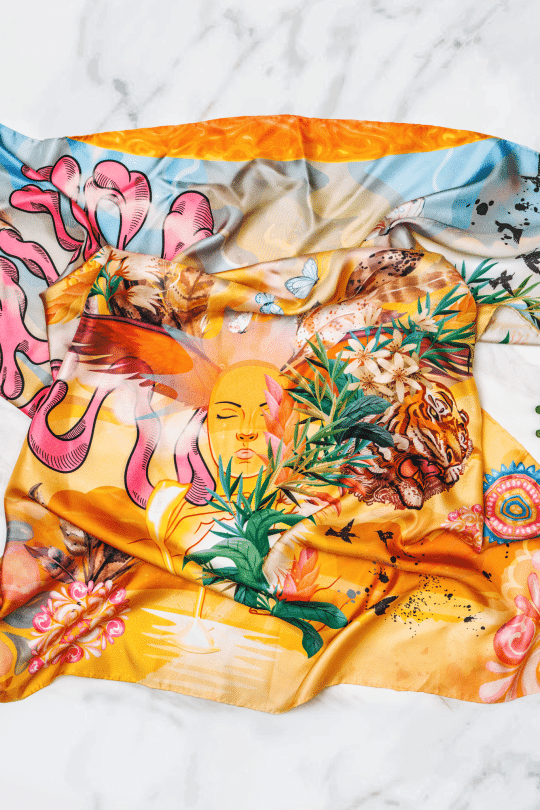 Das Seidentuch „Madre Medicina“ 85x85, geschmückt mit einer Reihe lebendiger Illustrationen wie Blumen, einem Tiger, einem heiteren Gesicht und abstrakten Designs, wird elegant auf einer weißen Marmoroberfläche präsentiert. Links liegt eine Sonnenbrille, während rechts eine Pflanze und eine Teetasse mit Untertasse stehen – was einen Hauch deutscher Mode-Eleganz verleiht.