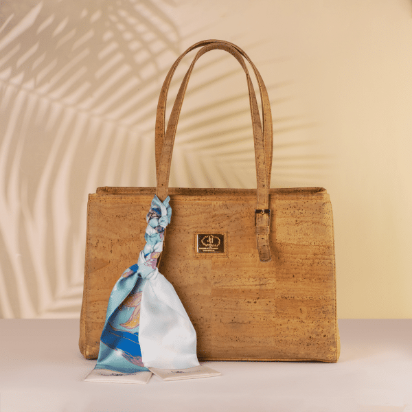 A beige handbag made of cork material with double handles and a gold logo plate on the front is adorned with the luxurious Seidentuch "Meerjungfrau," featuring shades of blue and pink, stylishly tied to one handle. The background showcases a subtle, soft palm leaf shadow.