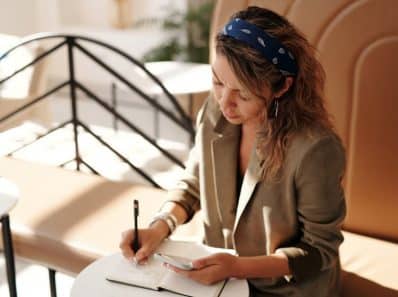 Eine Frau mit lockigem Haar, einem blauen Stirnband und einem Blazer sitzt auf einer Bank in einem sonnendurchfluteten Raum. Sie schreibt mit einem Stift in ein Notizbuch und hält in der anderen Hand ein Smartphone. Im Hintergrund sind ein geschwungenes Geländer und Tische zu sehen.