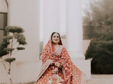 Eine Person in einem traditionellen rot-goldenen Hochzeitsoutfit steht im Freien. Sie trägt einen reich bestickten Lehenga mit passendem Dupatta. Im Hintergrund ist ein Gebäude mit hohen weißen Säulen und einem kleinen Baum zu sehen.