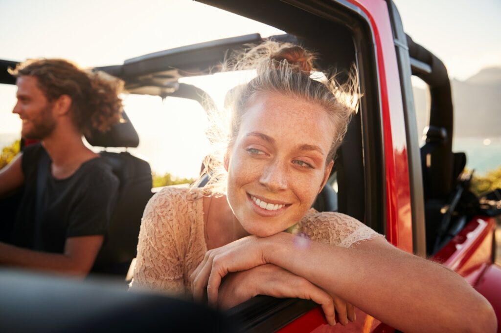 Millennial white couple on a road trip driving in open top car, women leaning on car door, close up