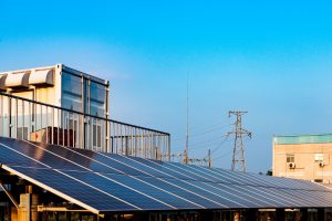 Vista di un impianto solare su un tetto, con pannelli fotovoltaici che catturano la luce solare, affiancato da linee elettriche e strutture industriali.