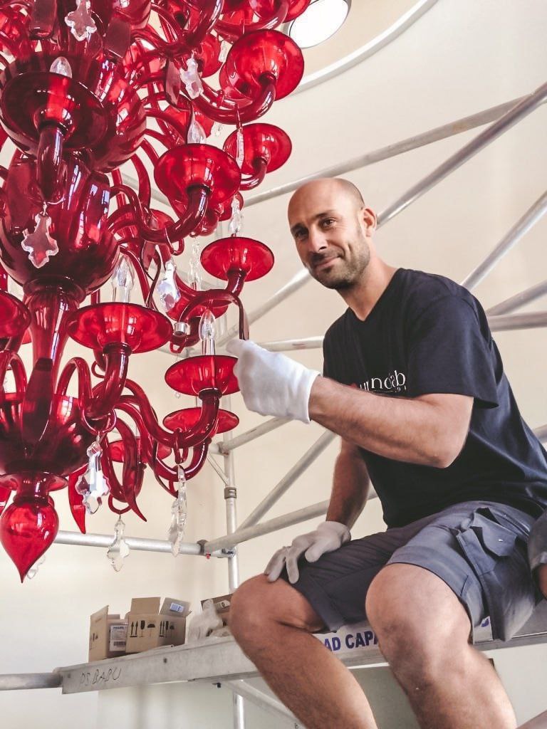 Nicola Polon, seduto su una piattaforma, sistema un lampadario di vetro rosso, indossando guanti per proteggere l'opera e lavorare con attenzione.
