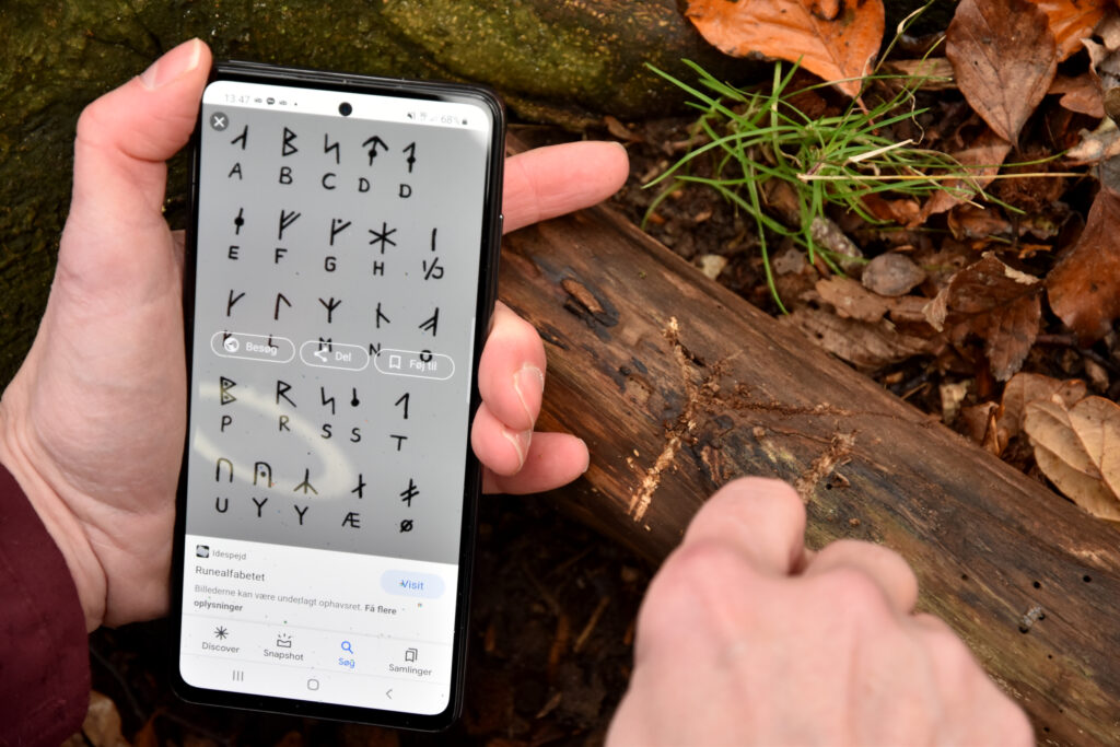 Mobiltelefon med runealfabet og en hånd der rister runer i et stykke træ.