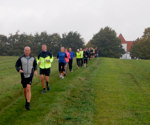 Løbere på en sti på en mark. I baggrunden, mellem nogle træer, en kirke.