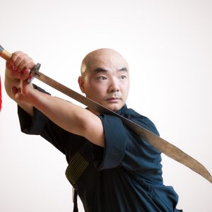 Shaolin-Meister Shi Yan Yao in schwarzer Trainingskleidung hält ein chinesisches Breitschwert mit beiden Händen in einer dynamischen Abwehrposition vor einem hellen, neutralen Hintergrund. Am Griff des Schwertes hängt ein auffälliges rotes Tuch.