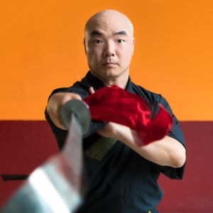 Porträt von Shaolin-Meister Shi Yan Yao im Fokus, der ein Kung-Fu-Breitschwert direkt in Richtung Kamera hält. Die Klinge im Vordergrund ist unscharf, während sein fokussierter Blick, die schwarze Trainingskleidung und ein rotes Schwerttuch vor einem orange-roten Hintergrund scharf abgebildet sind.