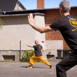 Perspektive über die Schulter eines Schülers auf Meister Shi Yan Yao, der im Innenhof des Shaolin Tempel Deutschland in Gollenberg einen tiefen Kung-Fu-Stand mit ausgestreckter Hand demonstriert.